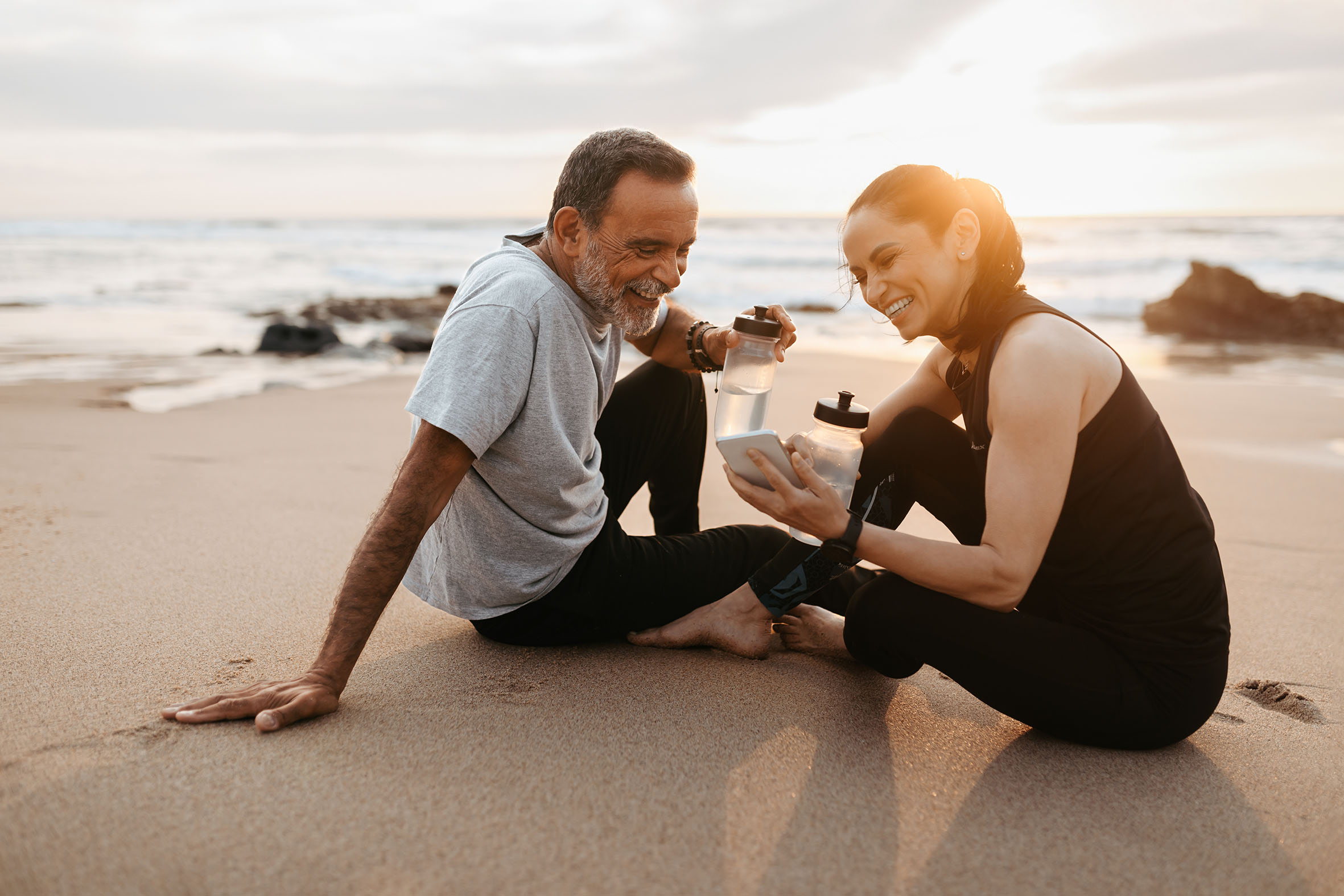 Happy mature caucasian man and lady in sportswear rest from fitness, sit on sand with bottles of water